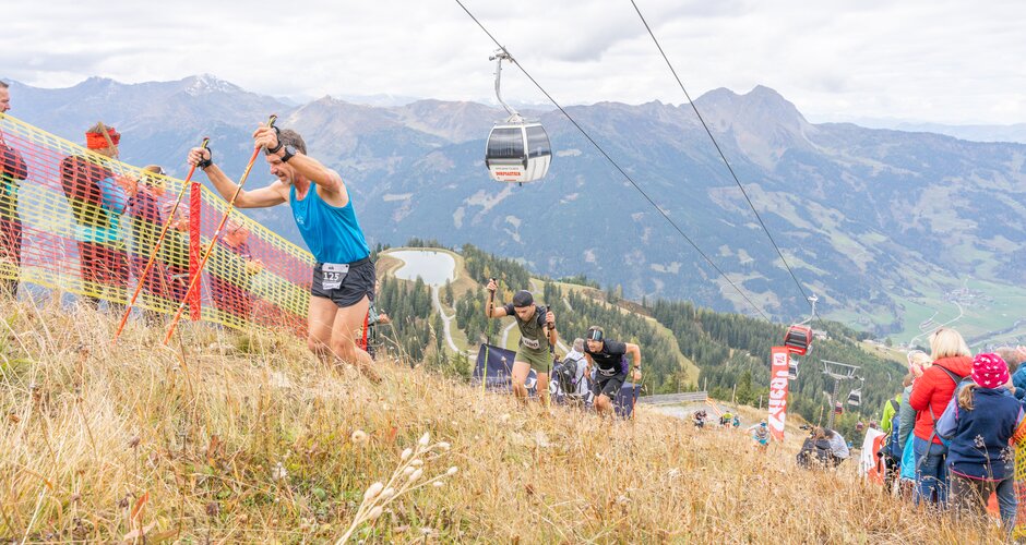 Beinhoat racers climb steep slope with poles, cheered by spectators; cable car and mountain scenery in the background. | © Beinhoat