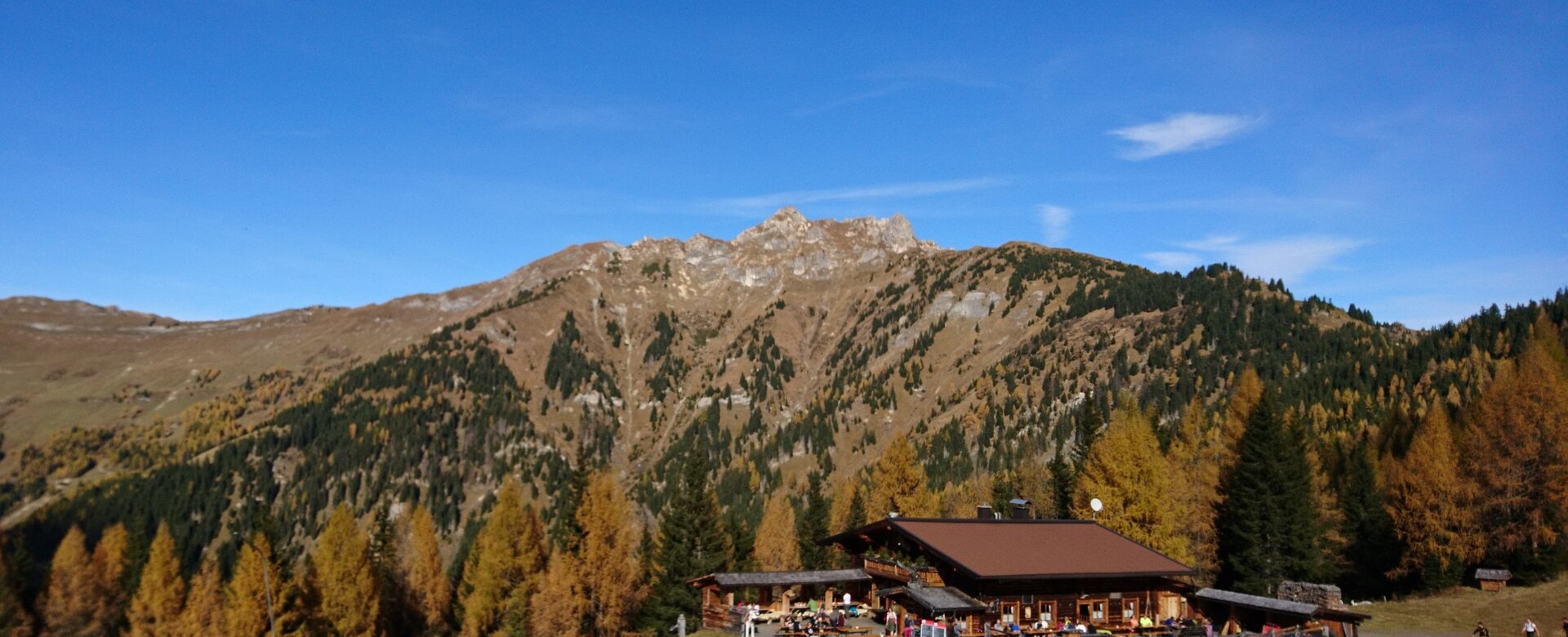 Holzhütte mit Sonnenterrasse im Herbstwald, Wanderer und Berge im Hintergrund