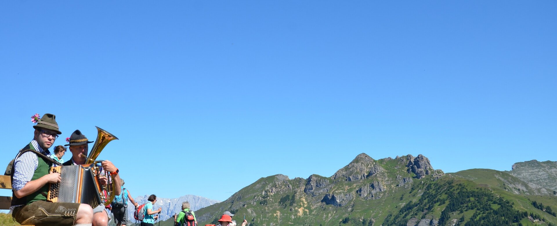 Zwei Musikanten in Tracht mit Tuba und Harmonika auf Wiese, Wanderer und Berge dahinter