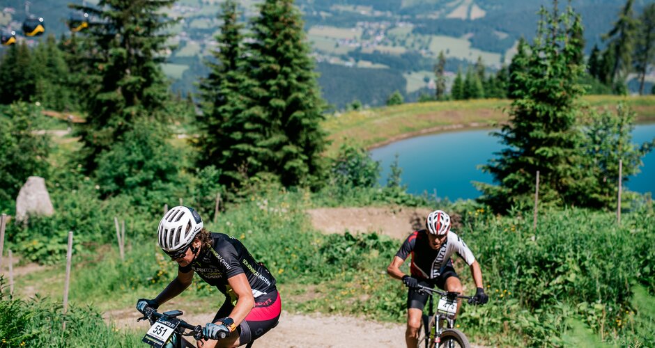 Zwei Mountainbiker fahren bergauf, im Hintergrund Gondeln und Bergpanorama bei der Alpentour Trophy in Schladming. | © Alexander Zauner