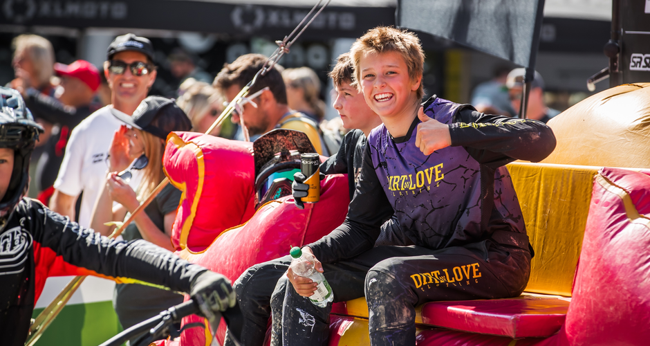 Young bikers sit in colorful event zone, one smiling and giving thumbs up. | © Sebastian Gruber
