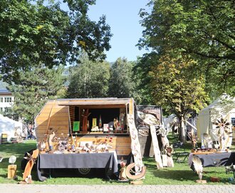 Market stall with wood art, metal decor and crafts in park, sunny day, white tents in background | © JOSalzburg