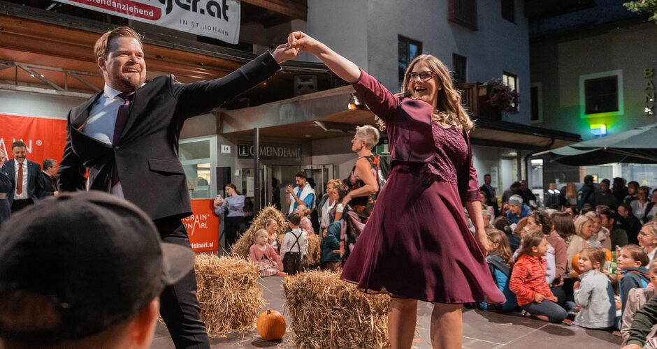 A man in a suit and a woman in a dress dance at a fashion show with straw bales and audience in the background. | © Belina Huttegger