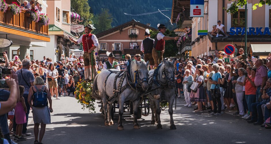 Two horses pull decorated carriage through village, surrounded by people at cattle drive. | © Lukas Anzinger