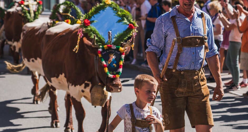 Man and boy in traditional dress lead decorated cows through a village at cattle drive. | © Lukas Anzinger