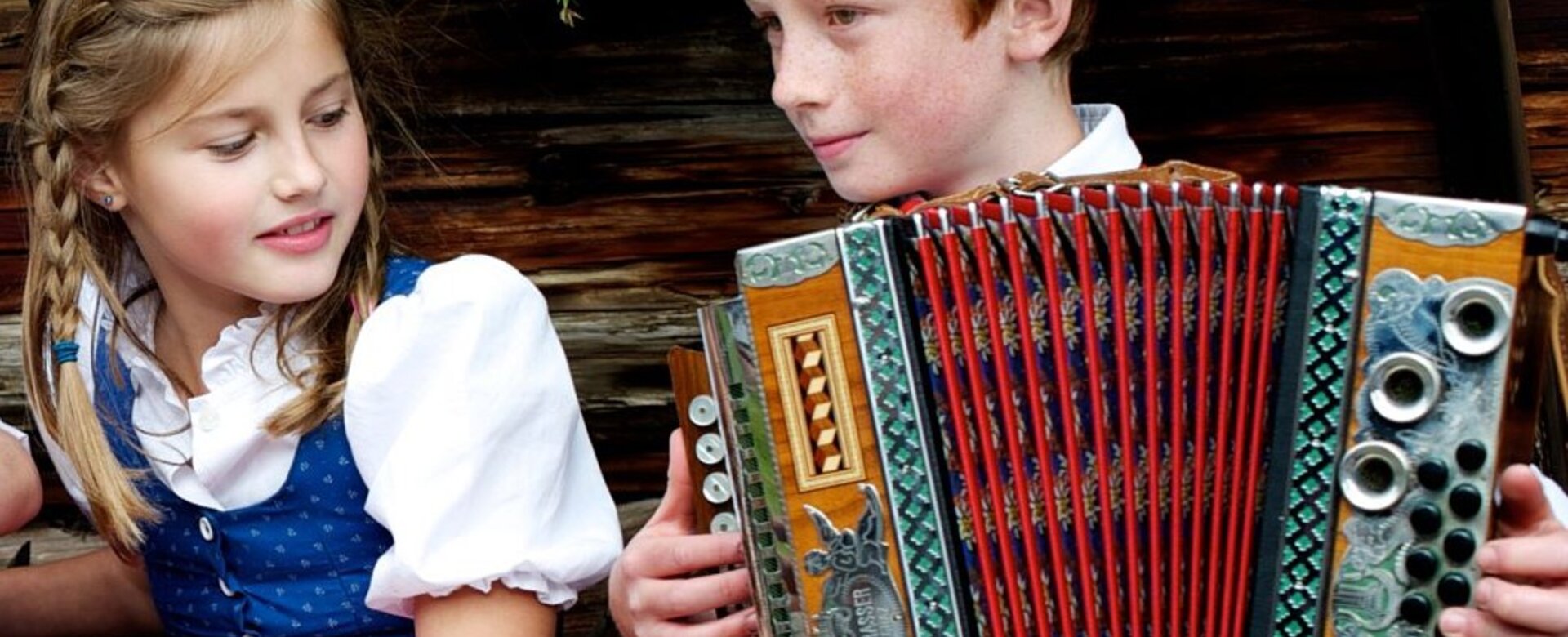 Girl in dirndl and boy in traditional clothes with accordion, wood wall and flowers | © Wagrain-Kleinarl Tourismus