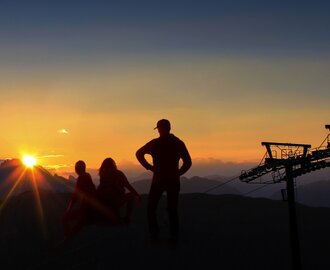 Three people in silhouette watch sunrise over mountains with cable car pylons.