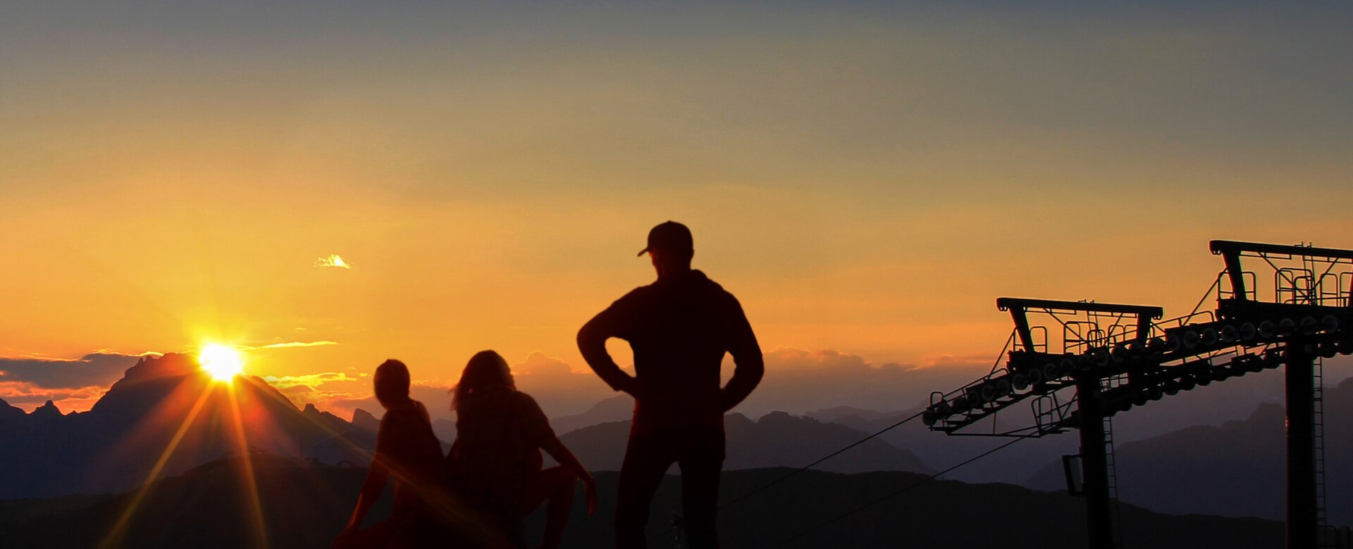 Three people in silhouette watch sunrise over mountains with cable car pylons.