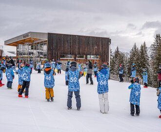 Zahlreiche Kinder in blauen Westen stehen im Schnee vor Gebäude beim Skicamp. | © Shuttleberg / Absolut Park