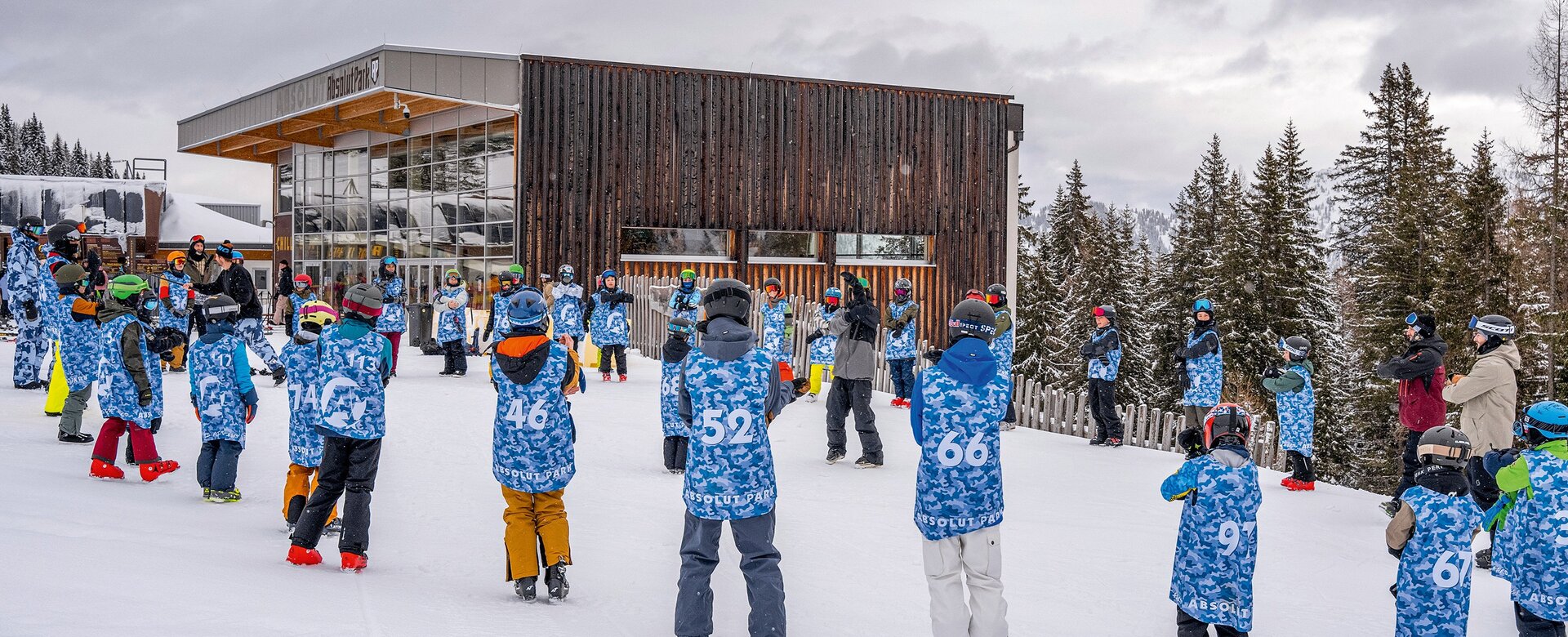 Many kids in blue vests standing in snow near lodge during ski camp session. | © Shuttleberg / Absolut Park