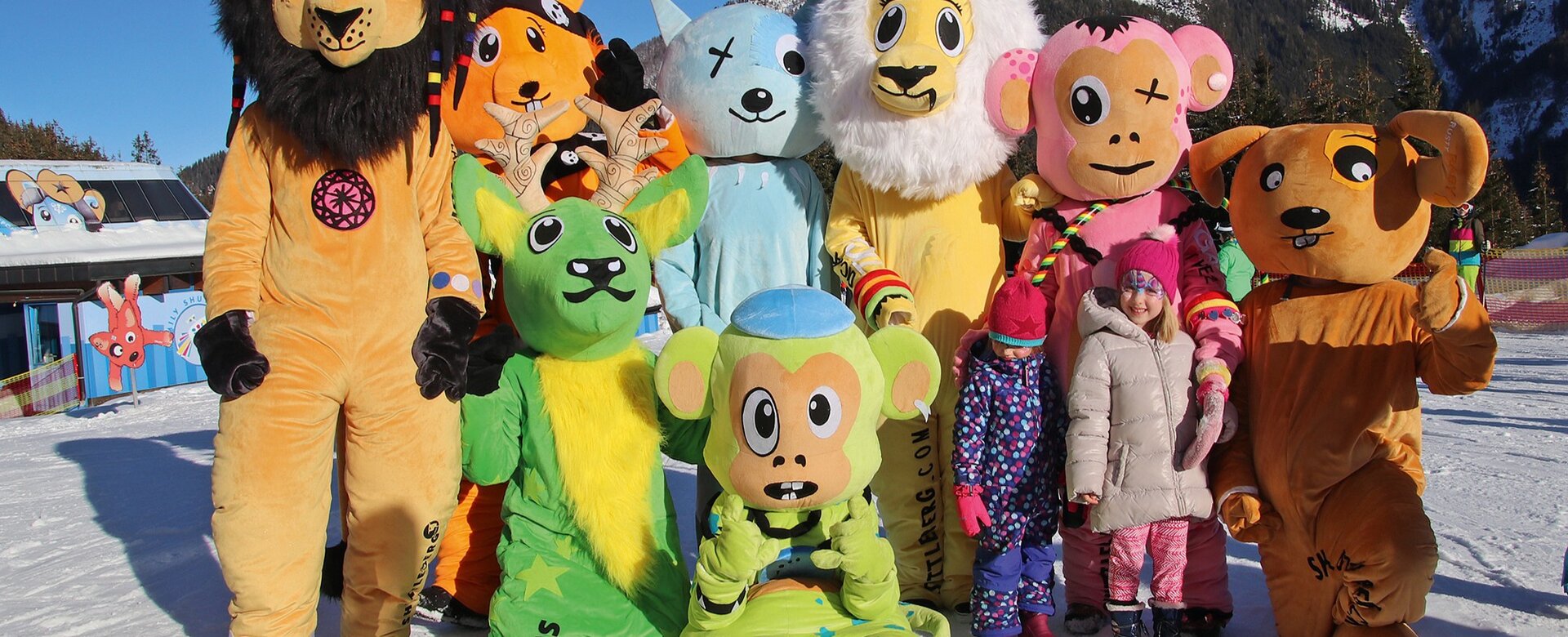 Zehn bunte Tiermaskottchen mit zwei Kindern im Schnee vor Bergkulisse.