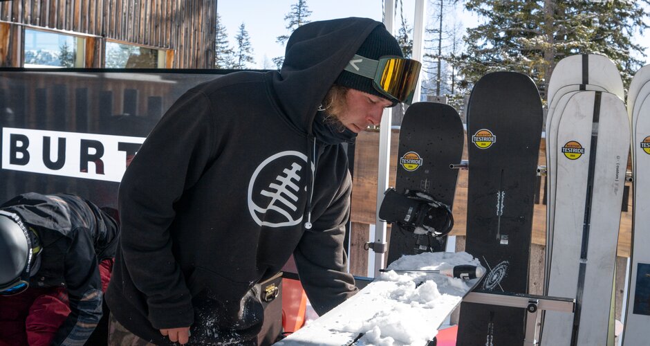 Person with black jumper and ski goggles on head makes adjustments to a white snowboard | © Juraj Opalek