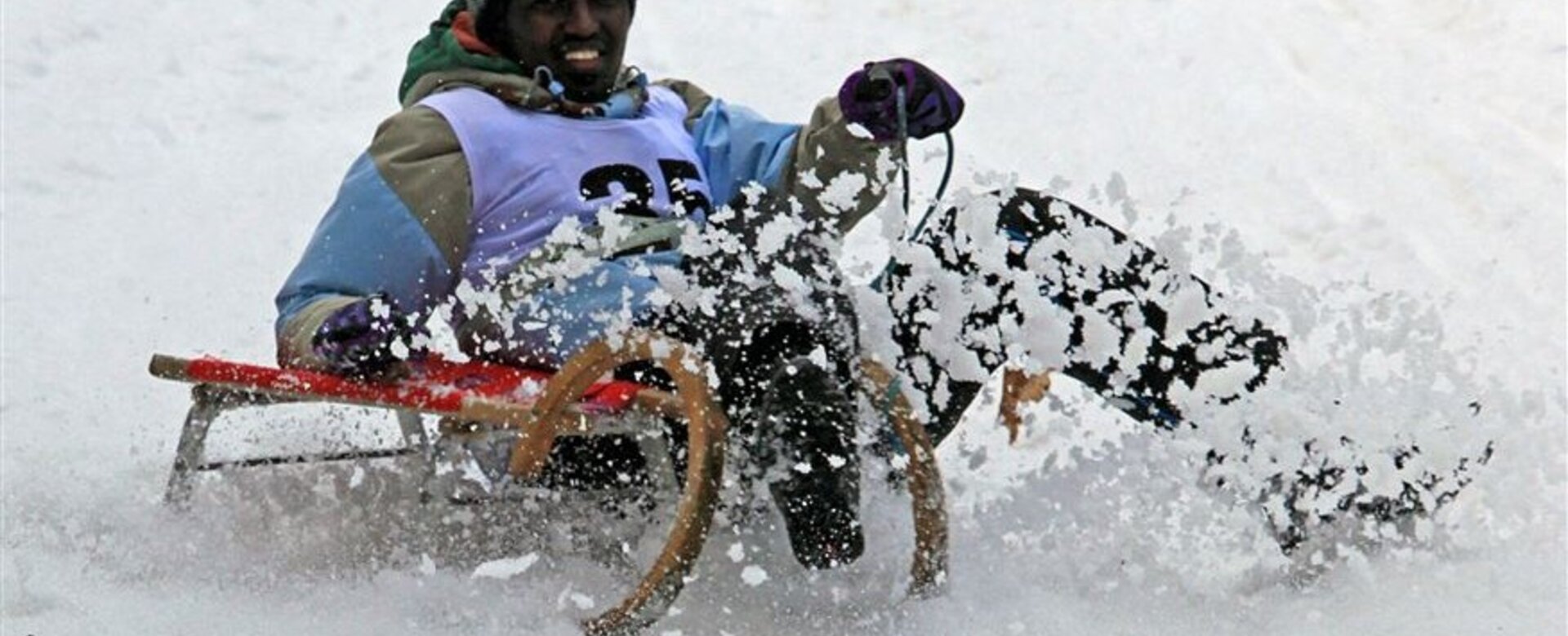 Ein Mann fährt mit Holzschlitten durch Schnee, aufgewirbelter Schnee im Vordergrund.