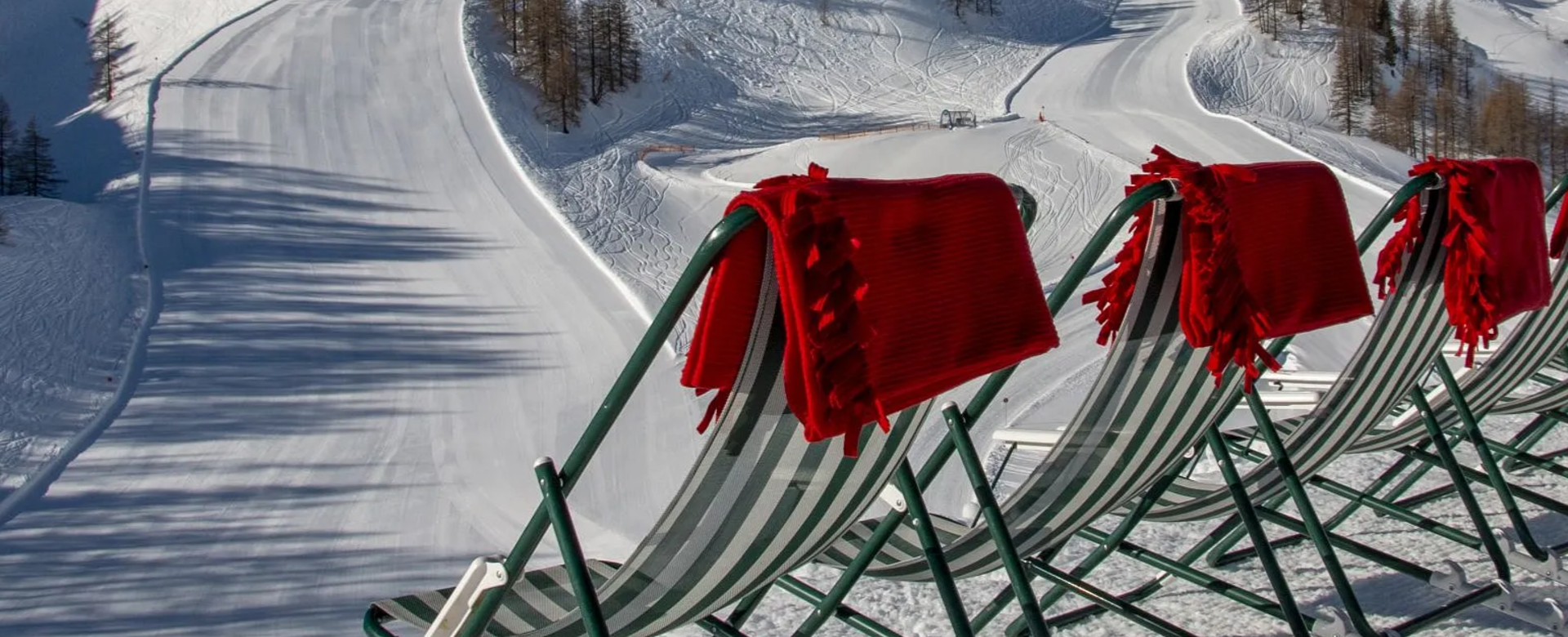 Deck chairs with red blankets on snow in front of snowy mountain landscape and ski slope