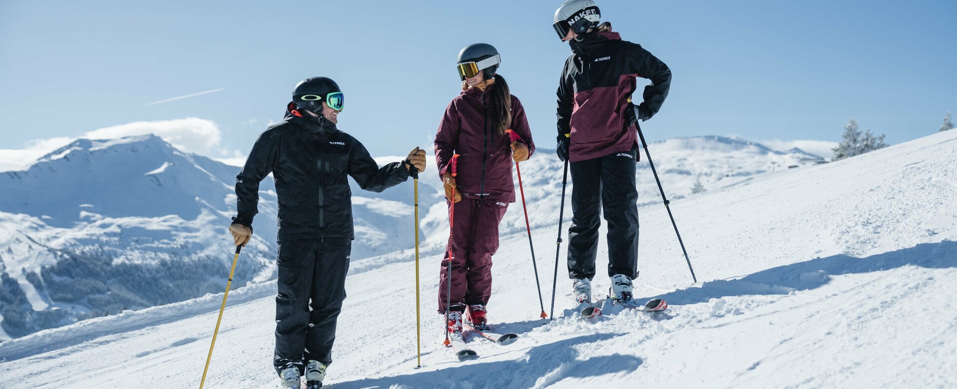 Three people in ski gear are standing on a slope talking with smiles and snow-capped mountains in the background | © Gasteinertal Tourismus GmbH, Christoph Oberschneider