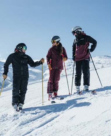 Three people in ski gear are standing on a slope talking with smiles and snow-capped mountains in the background | © Gasteinertal Tourismus GmbH, Christoph Oberschneider