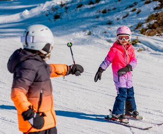 Two children in a ski course on a slope, one pointing at the other with a ski pole. | © Hochkönig Tourismus GmbH