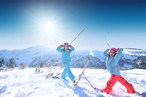 Two children are standing in deep snow, holding their ski poles to their helmets so that they look like horns, with their mouths wide open