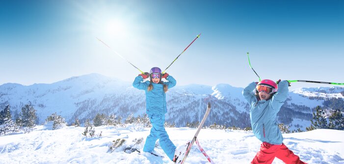 Zwei Kinder stehen im Tiefschnee und halten sich die Skistöcke an den Helm, so dass sie aussehen wie Hörner, und haben den Mund weit offen