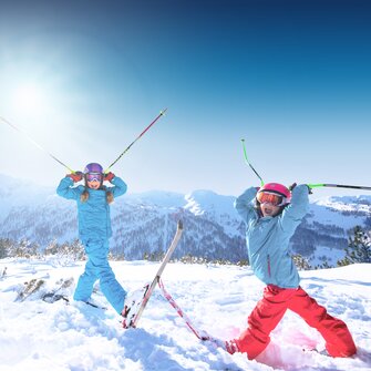 Two children are standing in deep snow, holding their ski poles to their helmets so that they look like horns, with their mouths wide open