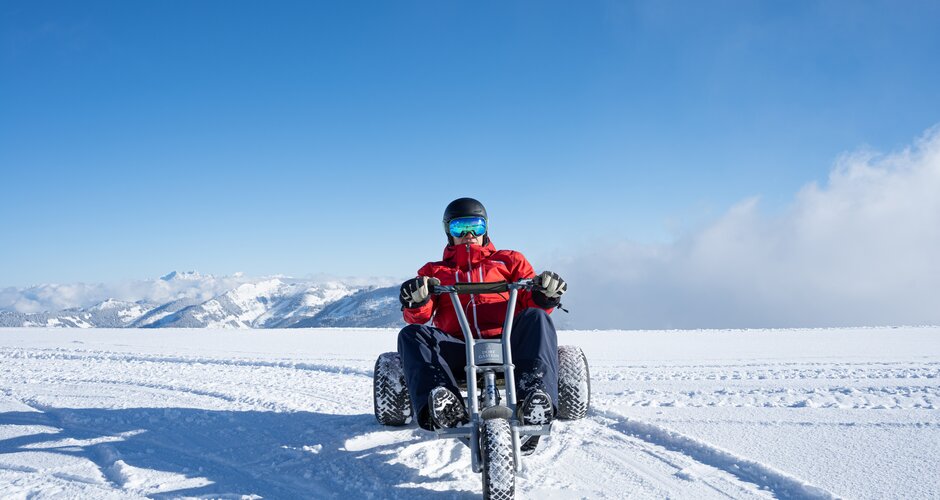 Person fährt mit Mountaincart über verschneites Hochplateau mit Alpenblick bei blauem Himmel in Dorfgastein | © Dorfgasteiner Bergbahnen