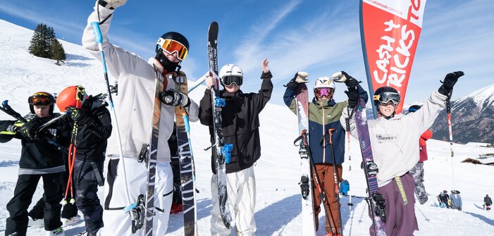 Four young freeskiers cheer in front of a Cash4Tricks flag, holding skis in the snow with blue sky and mountains behind them.