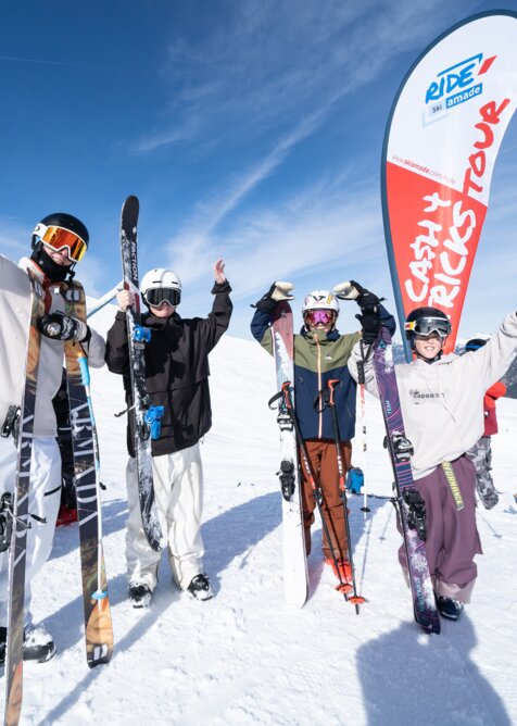 Four young freeskiers cheer in front of a Cash4Tricks flag, holding skis in the snow with blue sky and mountains behind them.