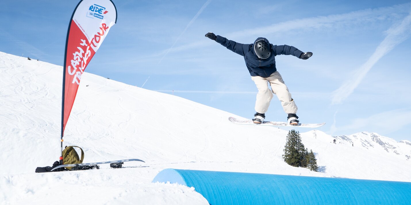 Ein Snowboarder springt bei Cash4Tricks über ein blaues Tube-Feature vor blauem Himmel, Eventflagge und Ausrüstung daneben.