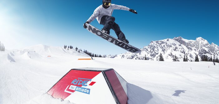 Snowboarder springt mit Grab über ein Obstacle im RIDE Ski amadé Snowpark, blauer Himmel und Berge im Hintergrund.