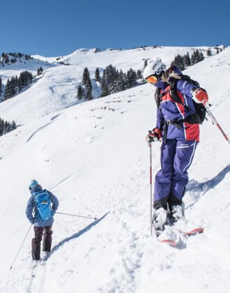 Zwei Freerider stehen leicht versetzt und mit dem Rücken zueinander auf einem Hang und sehen ins Tal hinunter