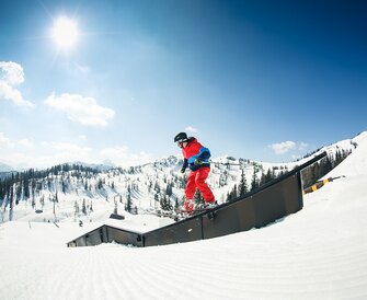 Freeskier in red-blue outfit slides on rail in snowpark with sunlight and snowy alpine scenery in background.
