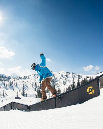Snowboarder in blauem Hoodie slidet bei Sonne über Rail im Snowpark, verschneite Berge und Wald im Hintergrund.