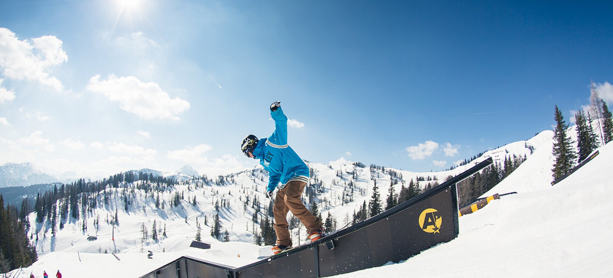 Snowboarder in blue hoodie slides on rail in sunny snowpark with snowy mountains and forest in the background.
