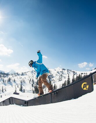 Snowboarder in blue hoodie slides on rail in sunny snowpark with snowy mountains and forest in the background.