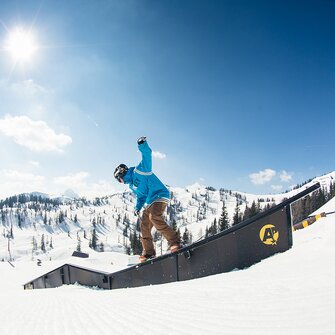 Snowboarder in blue hoodie slides on rail in sunny snowpark with snowy mountains and forest in the background.