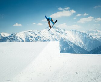 Snowboarder in blue hoodie does grab trick above kicker in snowpark with snowy Alps and bright sunshine behind.