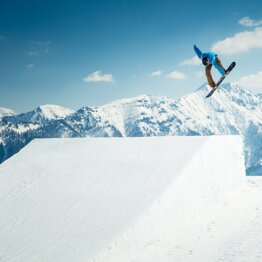 Snowboarder in blue hoodie does grab trick above kicker in snowpark with snowy Alps and bright sunshine behind.
