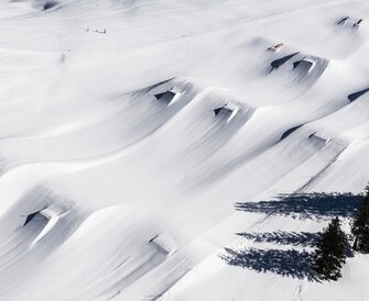 Aerial view of Kings Park Hochkönig showing fresh powder, shaped jumps, rails and distinct tree shadows on the snow.
