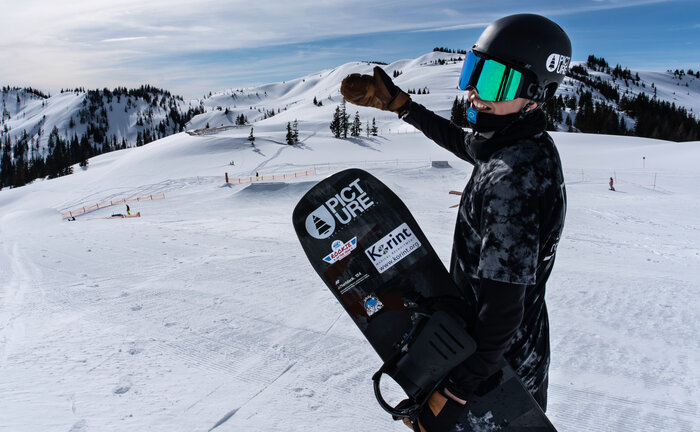 Smiling snowboarder in helmet and goggles points at Kings Park Hochkönig snowpark with obstacles in snowy terrain.