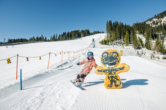 Kind mit Snowboard fährt bei Sonne auf der Funslope vorbei an bunter Figur im Skigebiet Shuttleberg | © Shuttleberg