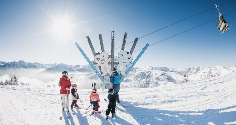 Kids with adult on skis at Family Run sculpture, chairlift and snowy mountains in the background | © Shuttleberg
