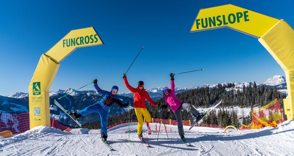 Three skiers in colorful outfits happily posing at the start of the Funslope and Funcross track in a sunny ski resort. | © Christine Höflehner