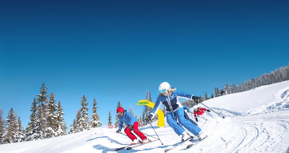 Two female skiers speed off from yellow Funcross gate with snow-covered trees and winter landscape in the background.