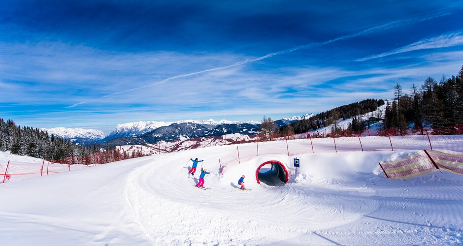 Drei Kinder auf Skiern fahren durch einen Schneetunnel auf der Funslope bei Sonne und mit Bergpanorama im Rücken.