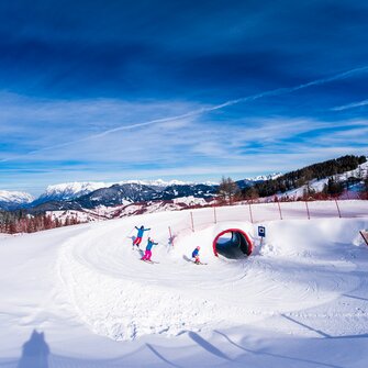 Drei Kinder auf Skiern fahren durch einen Schneetunnel auf der Funslope bei Sonne und mit Bergpanorama im Rücken.