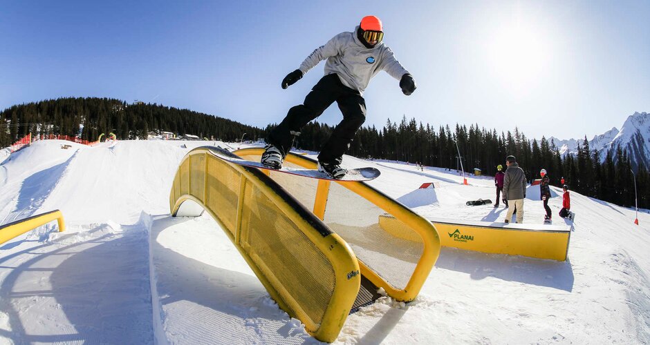 Snowboarder in a grey hoodie and orange beanie sliding on a yellow rail in Funpark Planai under bright sunshine. | © Roland Haschka QParks