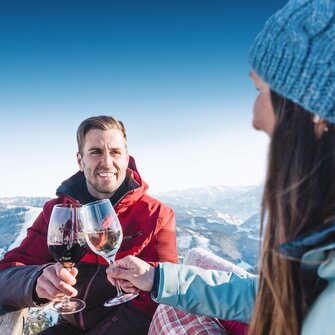 A man in a red ski jacket smiles and clinks glasses with a woman in a blue cap and blue ski jacket as they sit at a wooden table