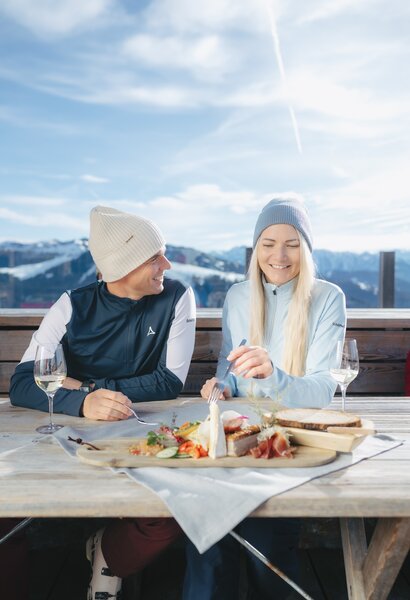 Zwei Personen essen auf Skihüttenterrasse Jause mit Käse, Wurst, Brot und Wein vor Bergkulisse | © Ski amadé