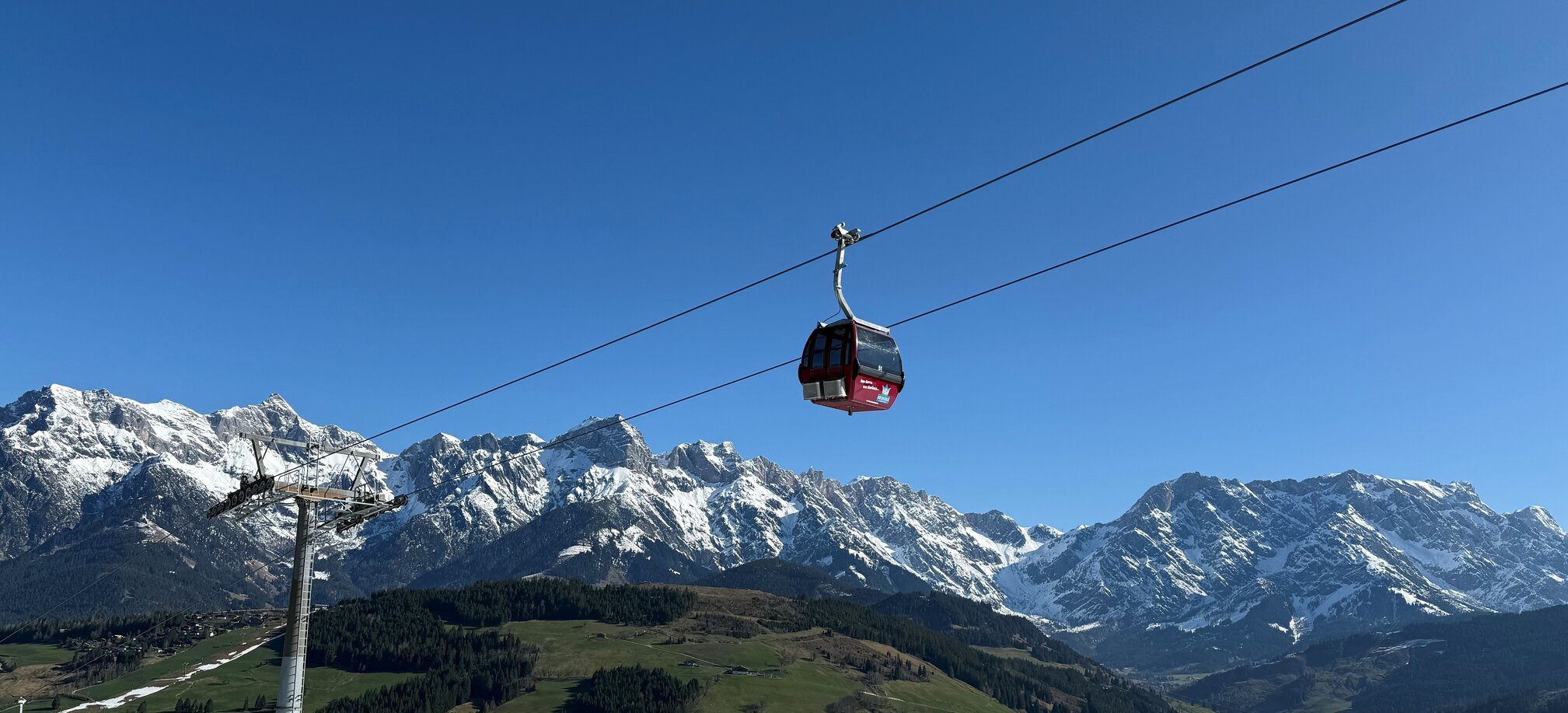 Rote Gondel der Abergbahn schwebt im Sommer vor grüner Landschaft und schneebedecktem Hochkönig-Massiv bei blauem Himmel. | © Aberg - Hinterthal - Bergbahnen AG