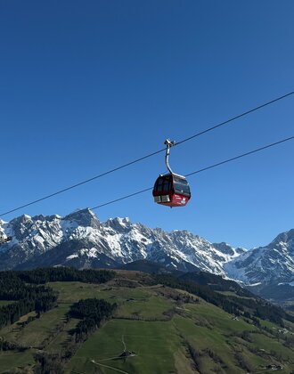 Red Abergbahn gondola glides in summer above green hills and snow-covered Hochkönig range under a clear blue sky. | © Aberg - Hinterthal - Bergbahnen AG